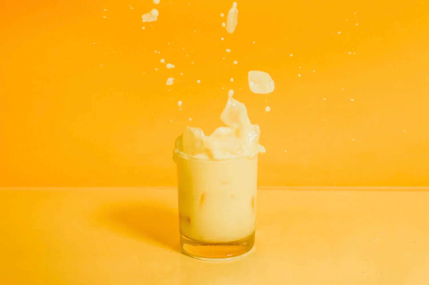 A glass of saffron latte made with The Fullest saffron latte mix, surrounded by splashes, on an orange background.