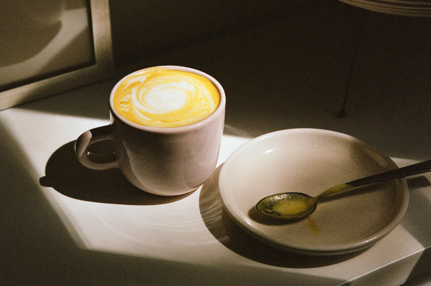 A warm and inviting saffron hot cocoa served in a ceramic mug, its golden swirl catching the sunlight on a minimalist tabletop. Beside the mug, a small white plate holds a spoon glistening with remnants of the saffron-infused drink.