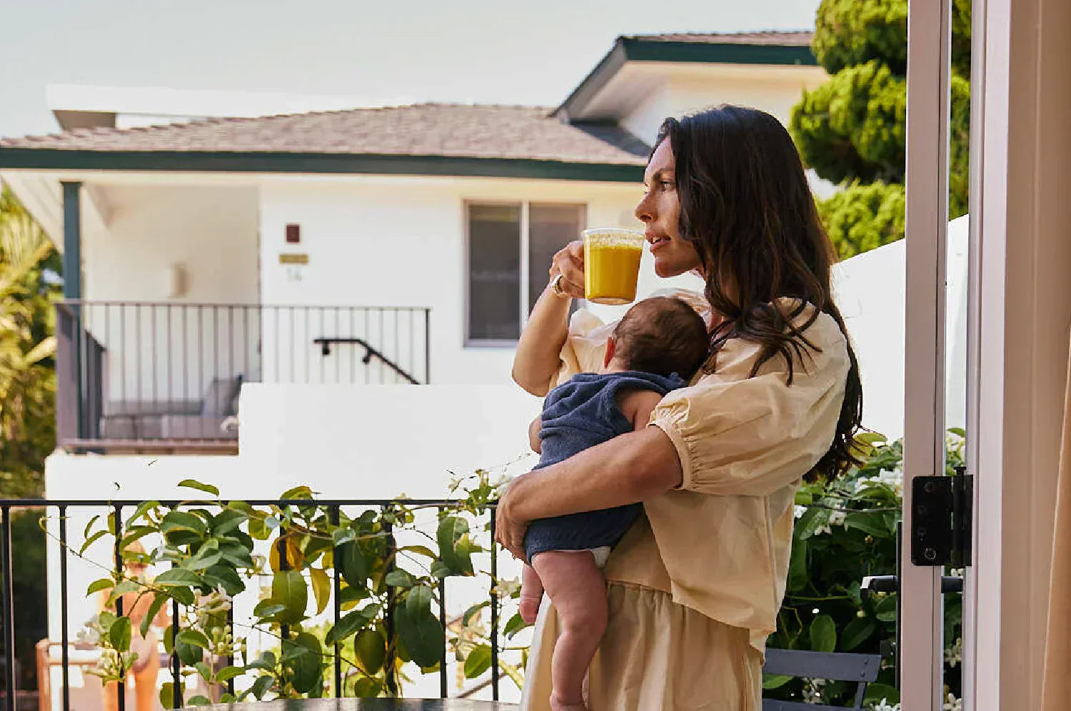 Woman sipping on a warm saffron latte standing on a balcony with her baby held close to her chest 