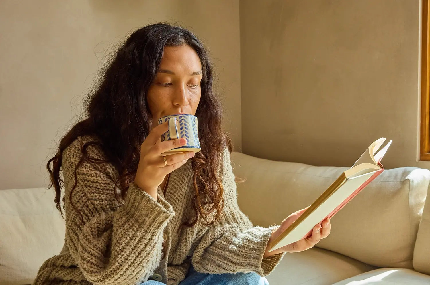High resolution of a woman sitting on a couch, reading a book in a serene state, holding a warm mug of a saffron latte with a neutral background 