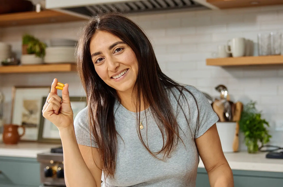 Woman smiling holding a saffron capsule in a warm light kitchen and open shelves with mugs, plants and cups 