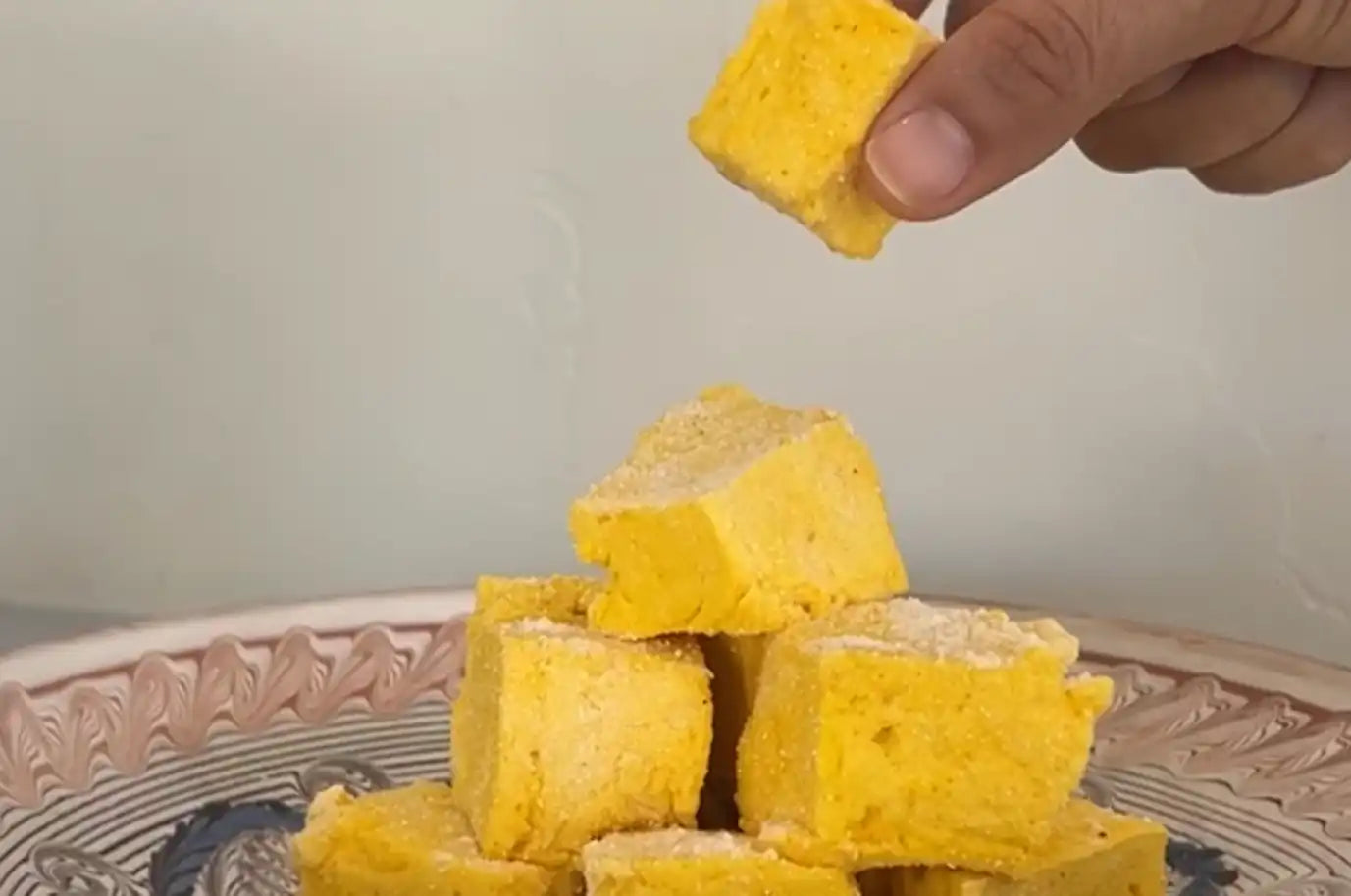 A hand picking up a cube-shaped, yellow saffron marshmallow from a decorative plate stacked with more of the same marshmallows. The marshmallows have a soft, fluffy texture and are lightly dusted with powdered sugar.