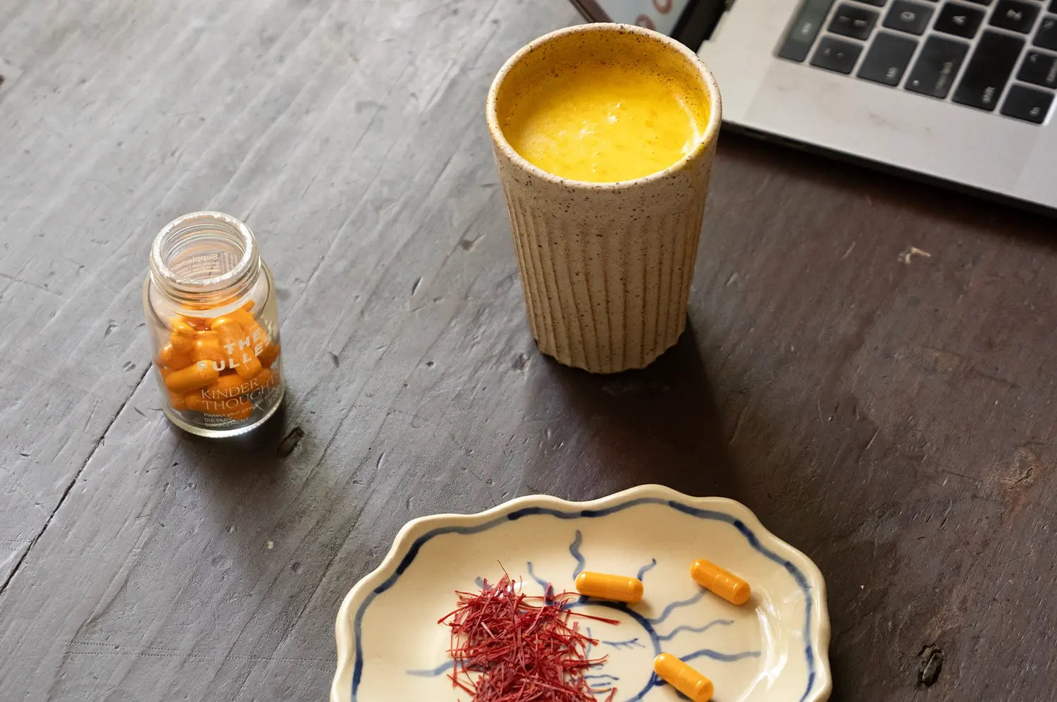 A cozy overhead shot of a warm saffron latte in a ceramic mug beside a dish of saffron threads, capsules and an open laptop —evoking a screen‑free summer ritual that nurtures eye wellness with natural glow