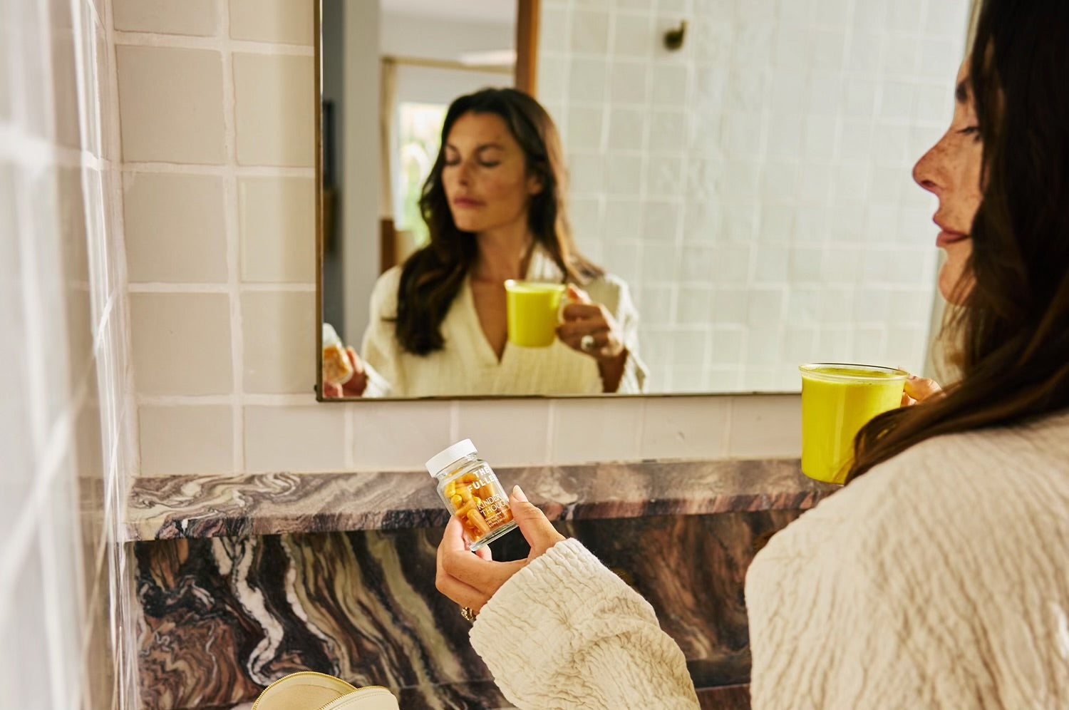 Model looking down at saffron capsules in one hand, saffron latte in the other with a granite counter and mirror reflection 