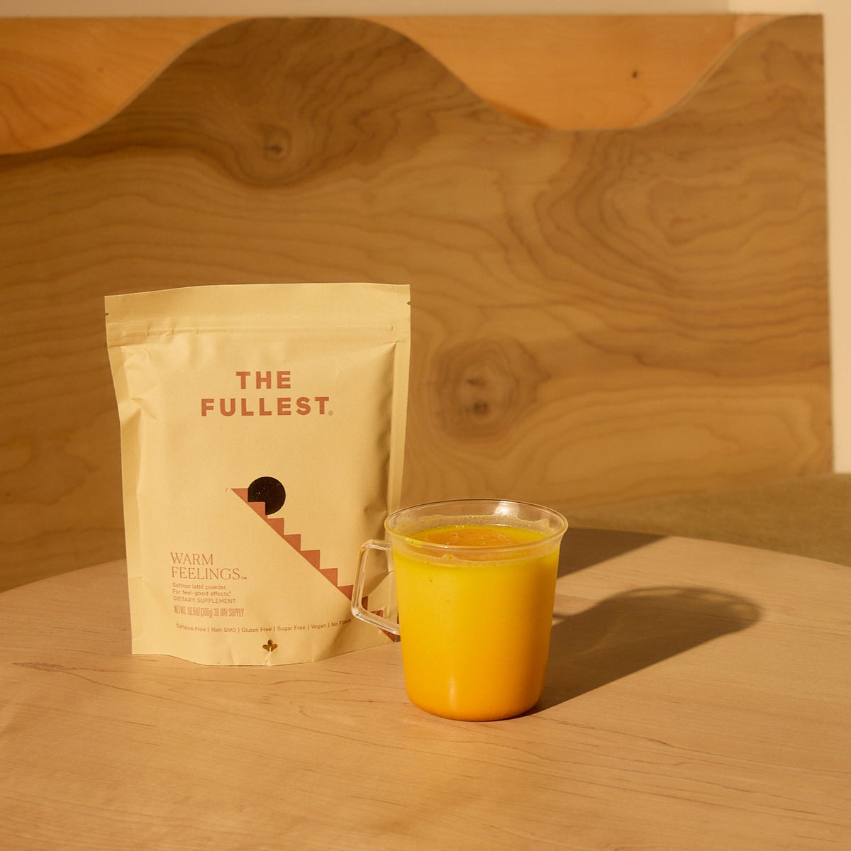 Yellow mug and &#39;The Fullest&#39; saffron latte package on a wooden table with a wooden bench in the background.