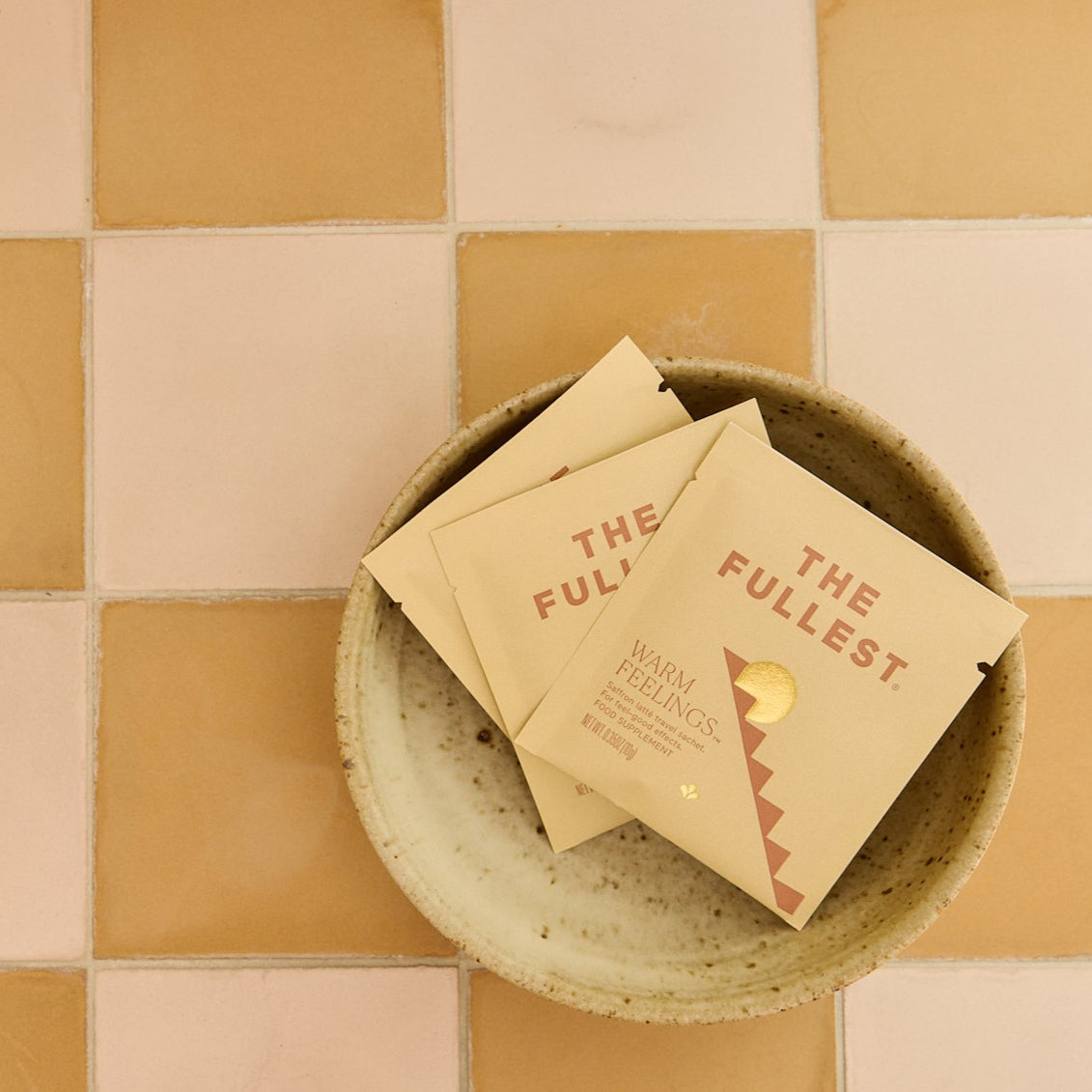 A ceramic bowl holds several beige THE FULLEST Saffron Latte Sachet 3-Day Supply packets on pink and tan tiles.