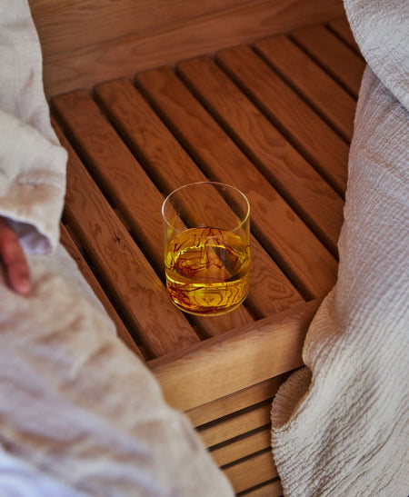 A clear glass tumbler containing saffron in liquid sits on a wooden slatted surface, surrounded by white textured fabric in a warm indoor setting.