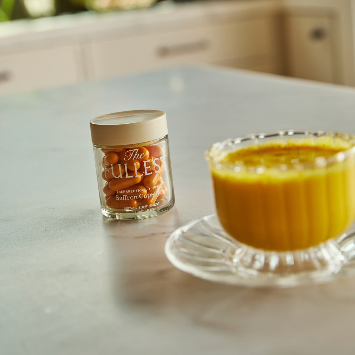 A glass jar of THE FULLEST Saffron Bundle for next to a cup of yellow saffron latte on a kitchen counter.