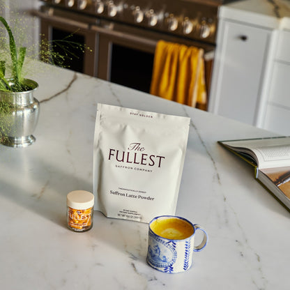 A Saffron Bundle featuring a white pouch of saffron latte powder, a small jar of saffron supplements, and a blue and white patterned mug filled with a yellow beverage on a marble countertop.