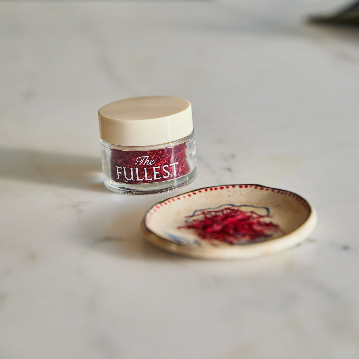 A small glass jar of THE FULLEST Saffron Threads next to a shallow decorative bowl containing a pinch of saffron.