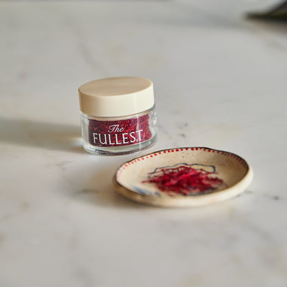 A small glass jar of THE FULLEST Saffron Threads next to a shallow decorative bowl containing a pinch of saffron.