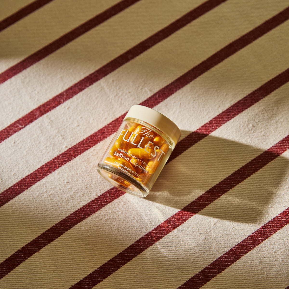 A small, clear glass jar filled with yellow THE FULLEST Saffron Capsules, sitting on a surface with brown and white stripes.
