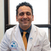 A man wearing a white medical coat with the name "Dr. Sadeghi" and a yellow patterned tie sits and smiles at the camera in an office setting.