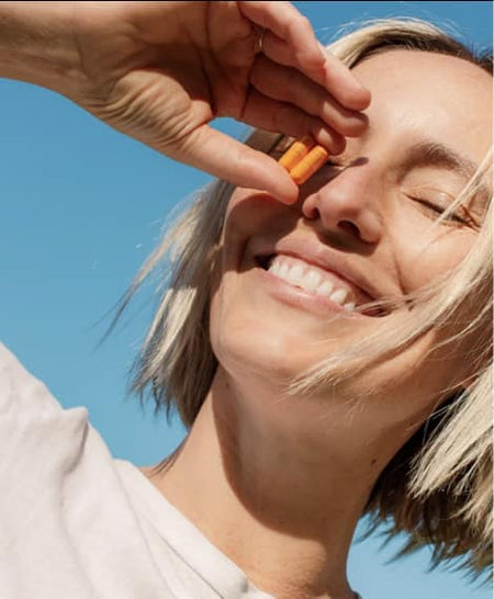 A smiling person with short blonde hair holds small carrot sticks over their nose, standing outdoors under a clear blue sky.