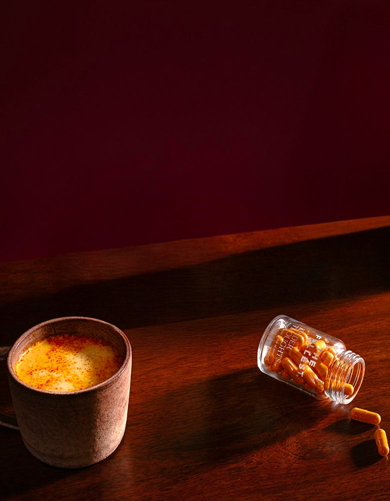 A ceramic mug filled with a yellow beverage sits on a wooden surface next to a tipped-over bottle spilling orange capsules.