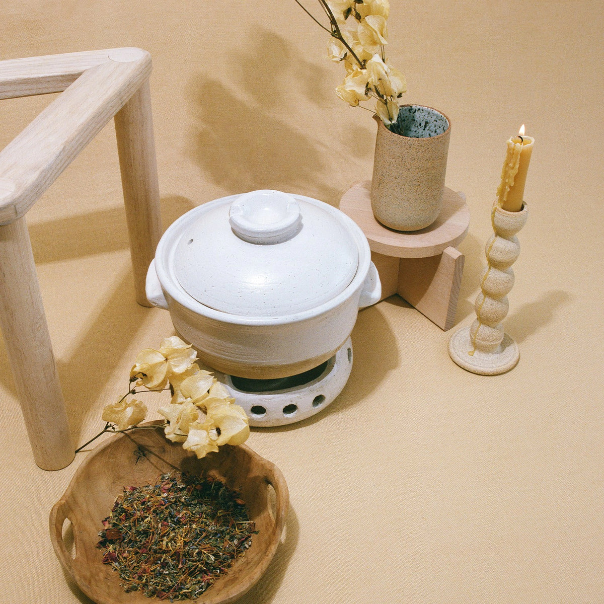 A cozy still life setup featuring THE FULLEST&#39;s Womb Protector Ritual Bundle on a warmer, a wooden bowl of dried herbs, a candle, and a vase with dried flowers on a beige surface.