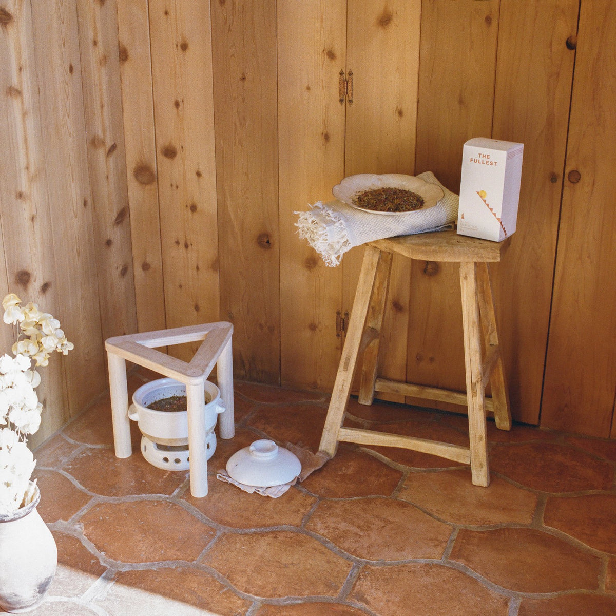 A cozy corner with a Womb Protector Ritual Bundle from THE FULLEST holding a book titled &quot;the poetry of r.&quot; and a bowl of pine cones, next to a small wooden chair with a white ceramic pot underneath.