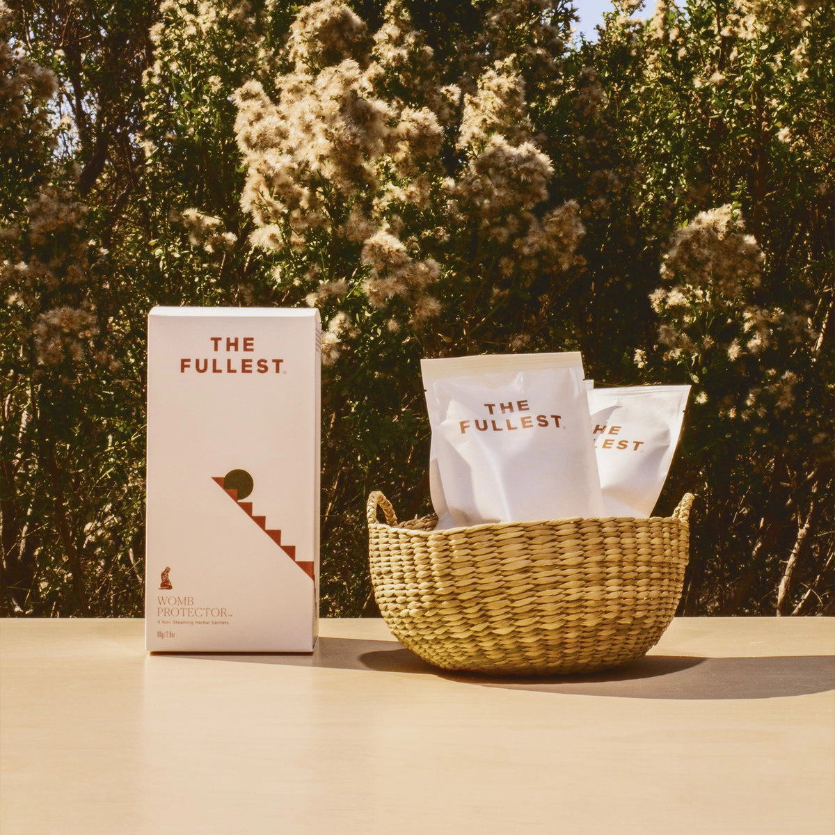Product packaging and sachets from THE FULLEST&#39;s Womb Protector Ritual Bundle displayed on a table with a wicker basket and a pelvic steaming stool, set against a backdrop of leafy shrubs and fluffy white flowers.