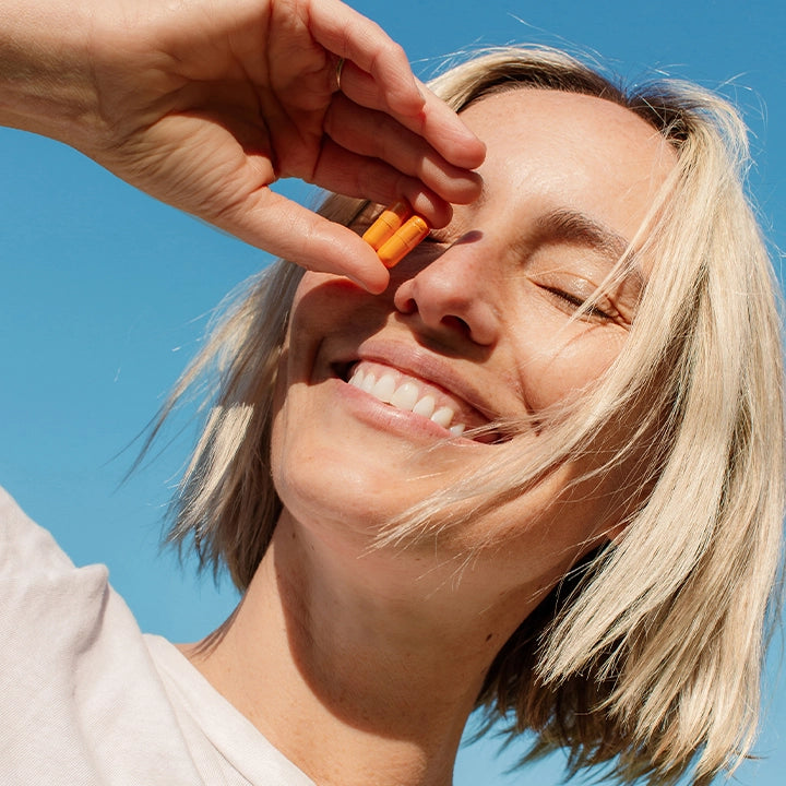 Smiling woman enjoying the sun while holding THE FULLEST's Kinder Thoughts saffron capsules, a natural supplement designed to support mood and well-being.