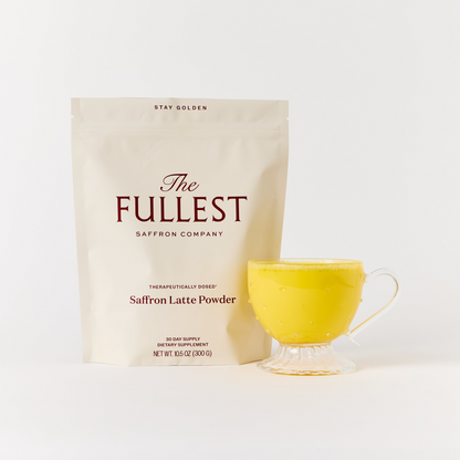 A package of THE FULLEST Saffron Latte 30-Day Supply is placed beside a clear glass cup containing a vibrant yellow, caffeine-free saffron latte that offers gentle mood support, all set against a plain white background.