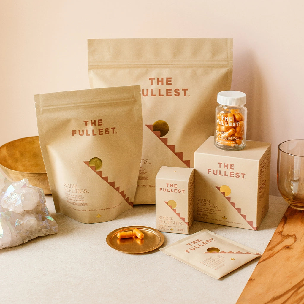 THE FULLEST's Warm Feelings Saffron Latte bulk, bag, and sachets, alongside their Kinder Thoughts capsules, on a kitchen table next to a crystal and glass.