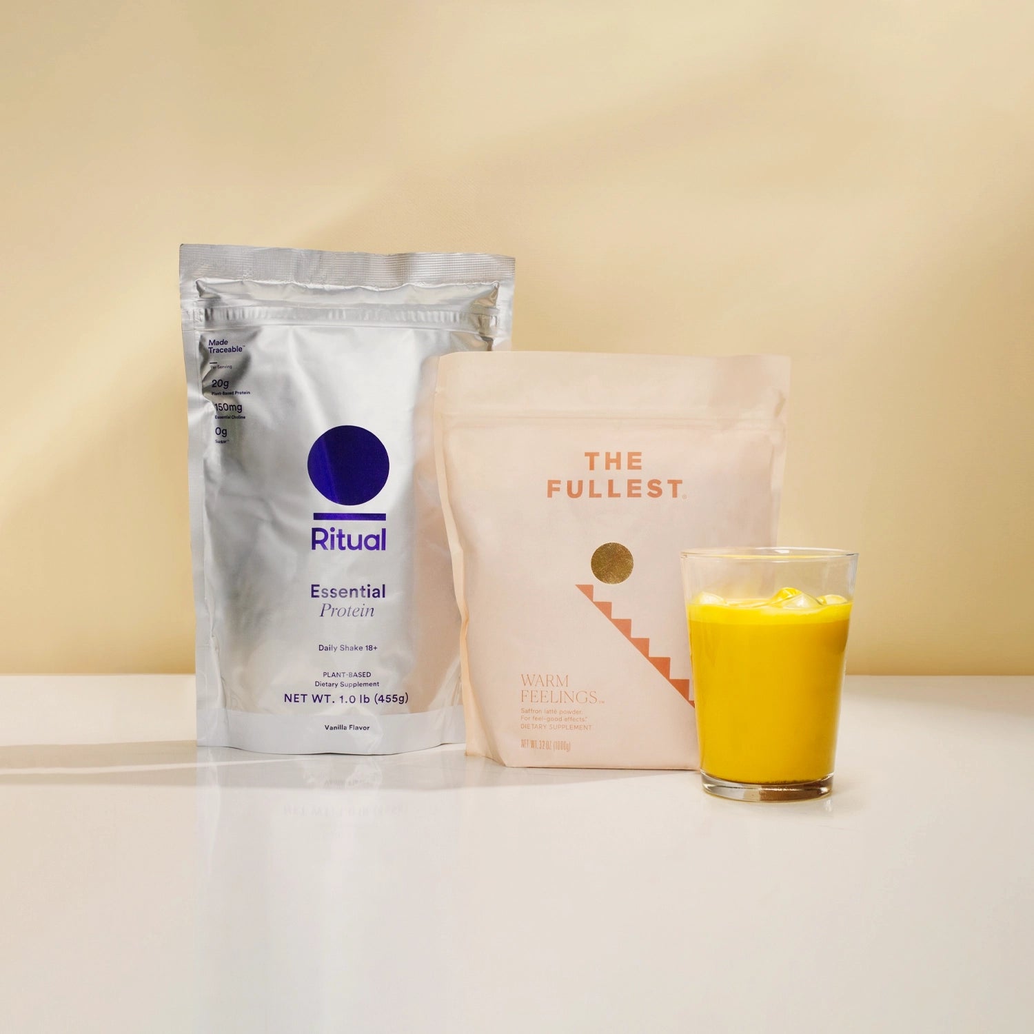 Two neatly standing pouches on a light surface against a soft yellow background. The left pouch is silver with a blue 'Ritual Essential Protein' label, and the right one is pink with 'THE FULLEST WARM FEELINGS' text.  Saffron beverage in front.
