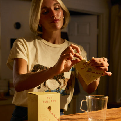 Someone opens a packet from THE FULLEST’s Saffron Latte 10 Single-Serving Sachets above a clear glass mug on a wooden countertop.