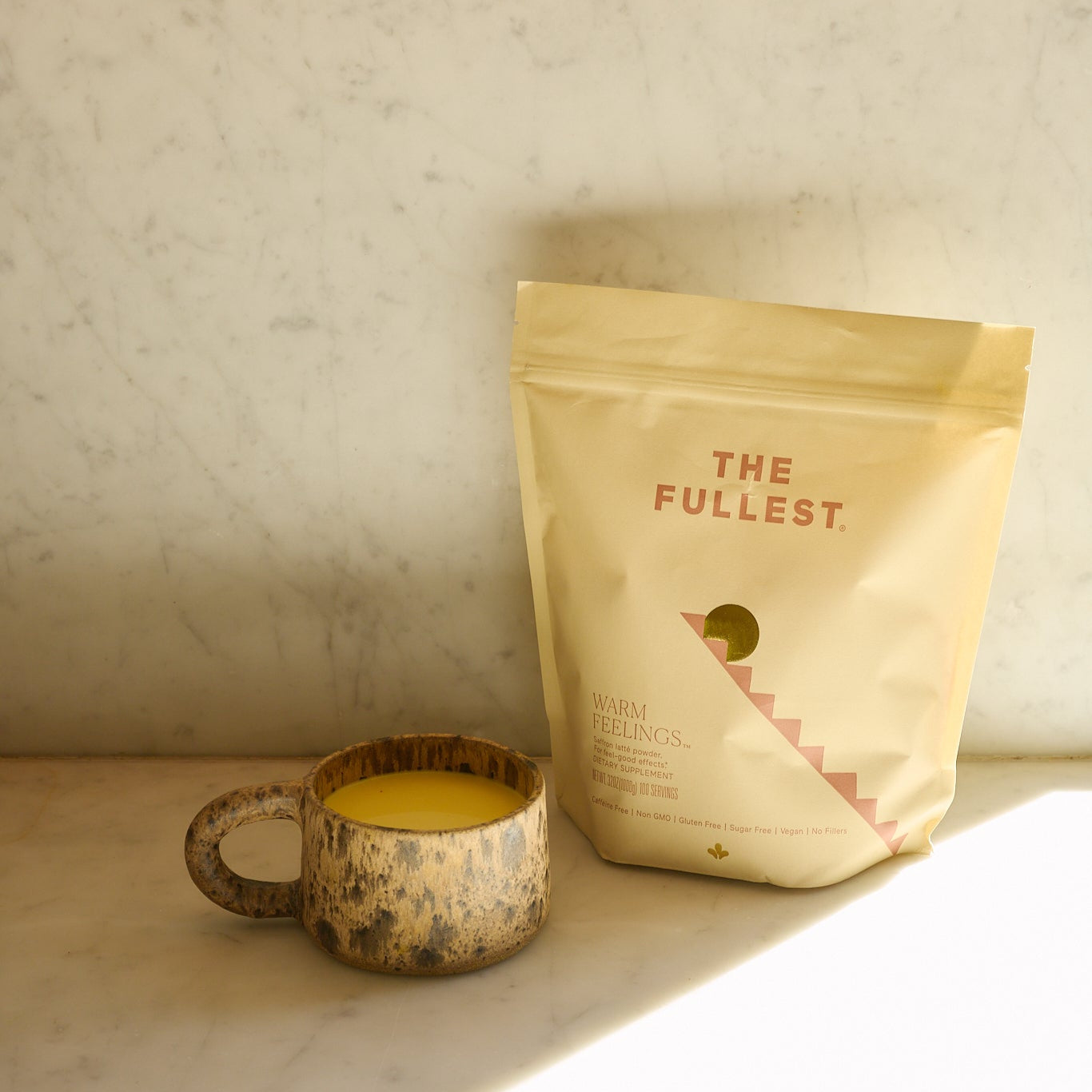 A rustic mug of yellow Saffron Latte sits beside a beige pouch labeled "THE FULLEST Saffron Latte 100-Day Supply" on a marble surface, sunlight casting gentle shadows.