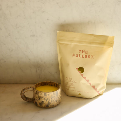 A rustic mug of yellow Saffron Latte sits beside a beige pouch labeled "THE FULLEST Saffron Latte 100-Day Supply" on a marble surface, sunlight casting gentle shadows.