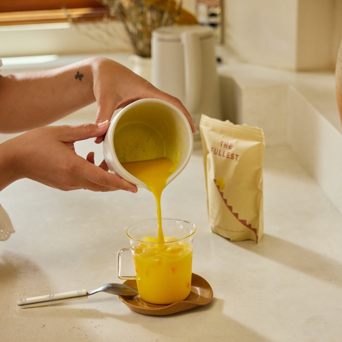 Someone pours a yellow, caffeine-free Saffron Latte from THE FULLEST from a white pitcher into a glass mug on a saucer, with a THE FULLEST packet in the background.