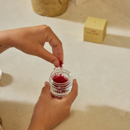 Hands place Saffron Threads into a glass jar on a light countertop, with a yellow box labeled "THE FULLEST" in the background, highlighting this premium product.