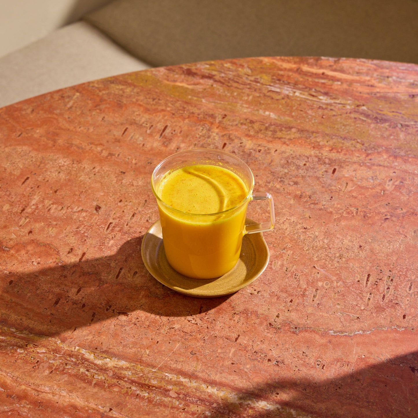 A clear glass cup of THE FULLEST Saffron Latte sits on a ceramic saucer on a round pink marble table in sunlight.