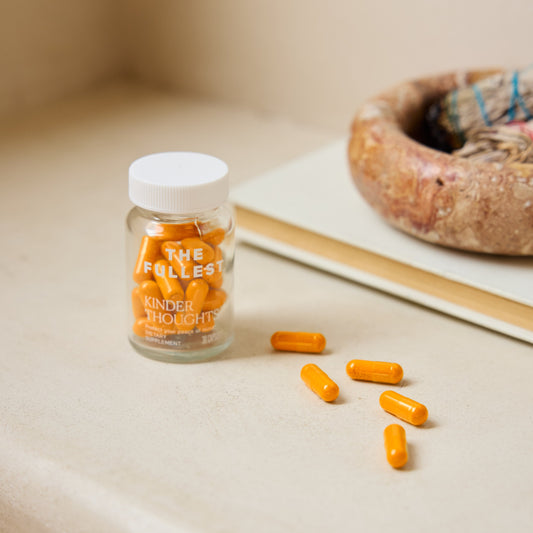 A clear jar labeled "THE FULLEST" Saffron Capsules sits on a beige surface, with six bright orange saffron capsules scattered beside it.