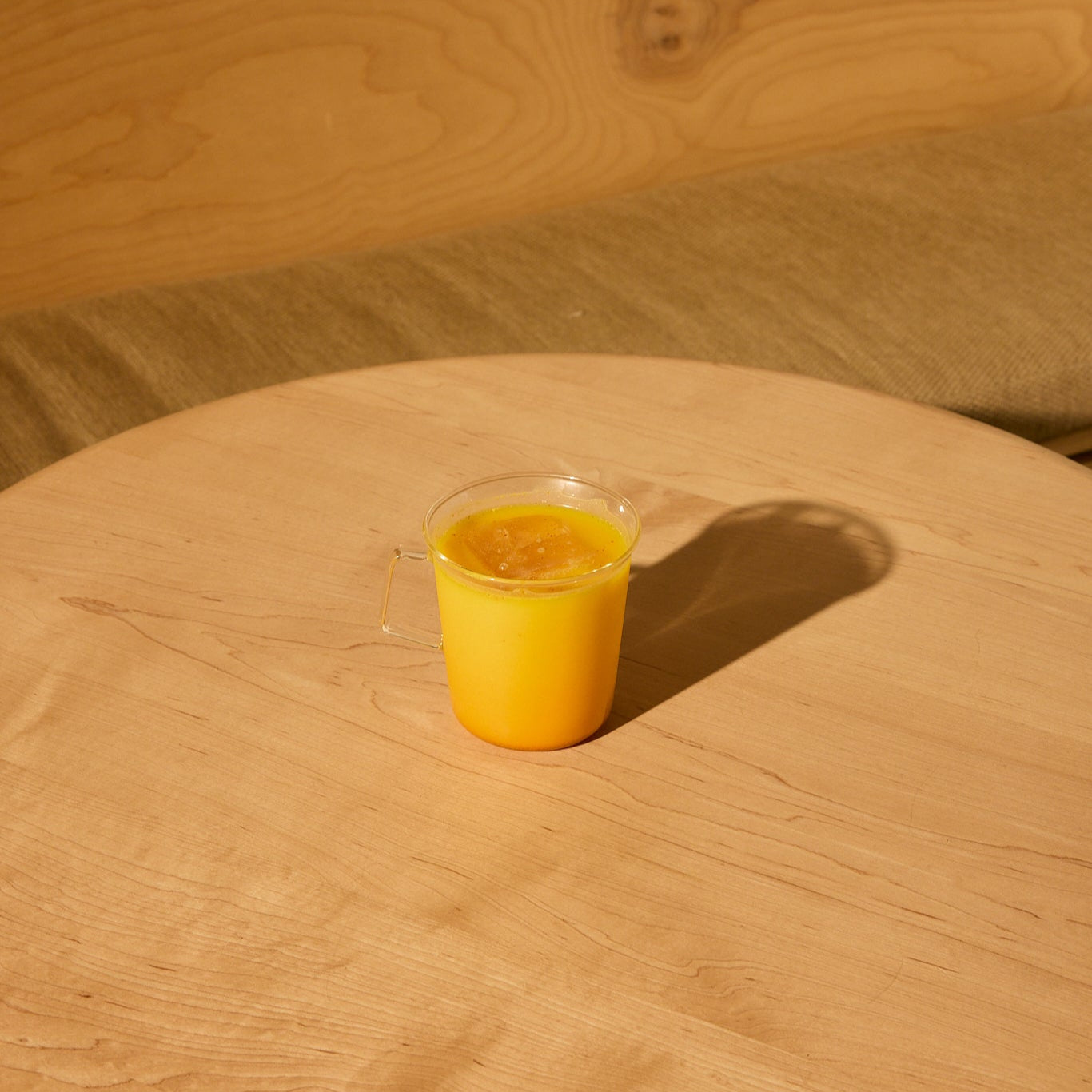 A clear cup of yellow-orange Saffron Latte, made with THE FULLEST’s Saffron Latte 10 Single-Serving Sachets, sits on a light wooden round table with a green cushioned bench and wood panel background.