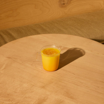 A clear cup of yellow-orange Saffron Latte, made with THE FULLEST’s Saffron Latte 10 Single-Serving Sachets, sits on a light wooden round table with a green cushioned bench and wood panel background.