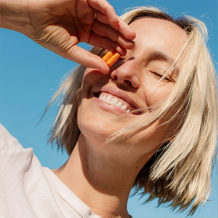 A woman with short blonde hair holding THE FULLEST Kinder Thoughts saffron capsules in front of her face.