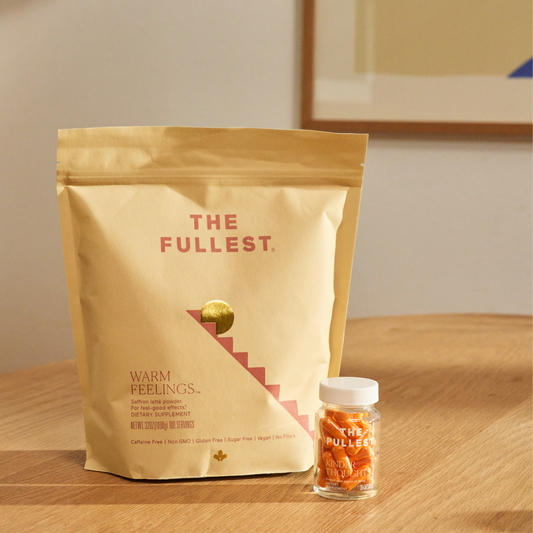 A beige pouch labeled "THE FULLEST." and a small clear bottle of orange-colored saffron capsules rest on a wooden table in a softly lit room.