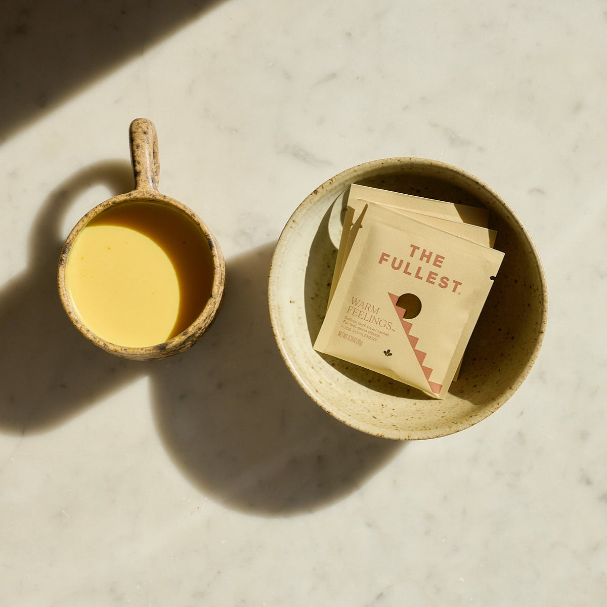 Two ceramic containers with saffron latte and packets labeled &#39;The Fulllest&#39; on a light surface.