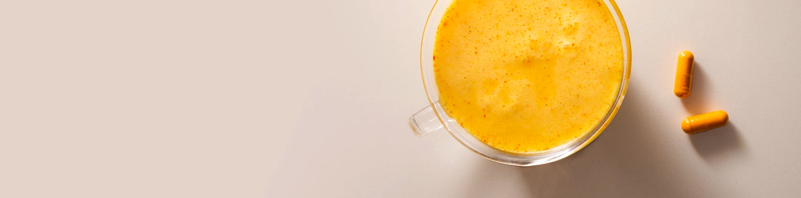 A clear glass mug filled with a vibrant yellow Warm Feelings saffron latte sits on a light beige surface, accompanied by two Kinder Thoughts saffron capsules placed to the right of the mug.