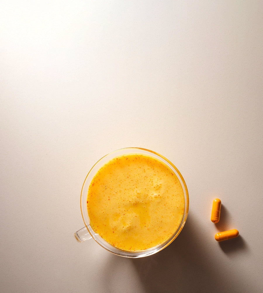 A clear glass mug filled with a vibrant yellow Warm Feelings saffron latte sits on a light beige surface, accompanied by two Kinder Thoughts saffron capsules placed to the right of the mug.