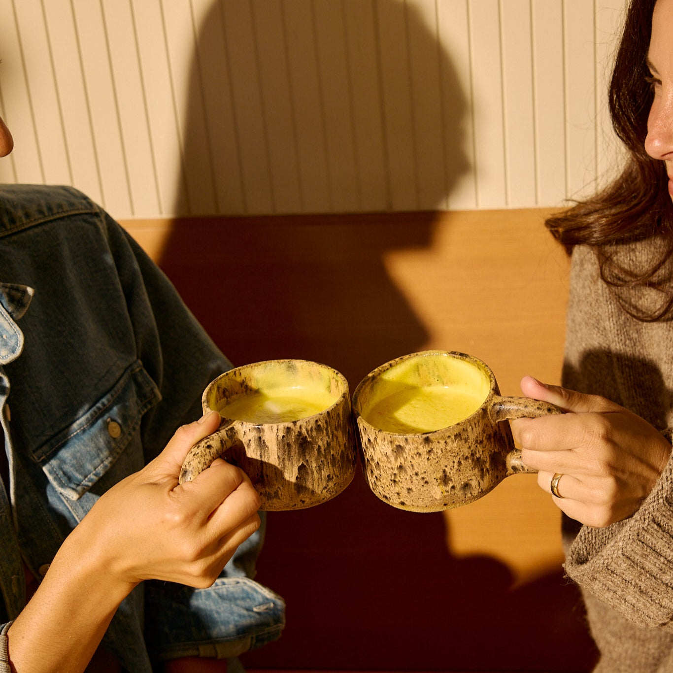 Two people sit together, clinking THE FULLEST Saffron Latte 100-Day Supply mugs filled with golden saffron lattes as sunlight casts shadows on the wall behind them.