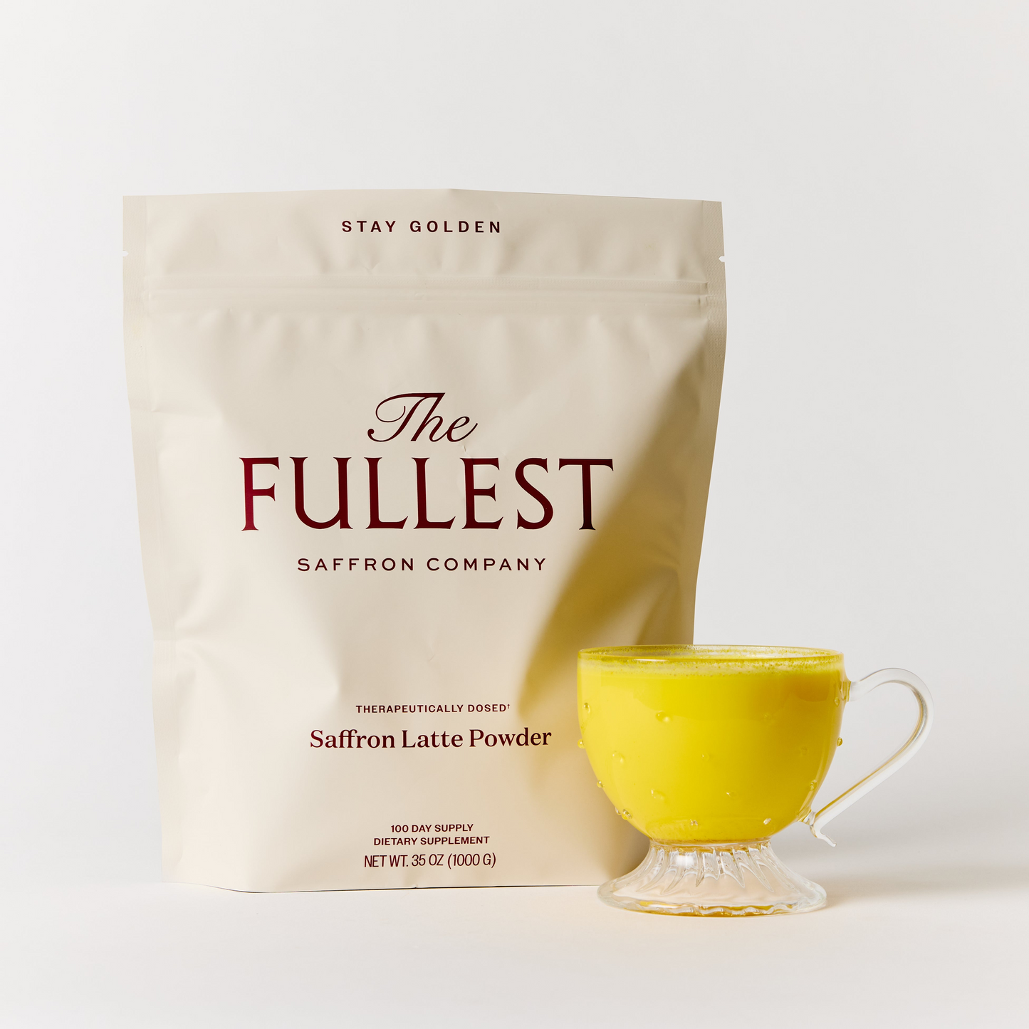A cream-colored bag labeled “Saffron Latte 100-Day Supply” from THE FULLEST stands behind a clear cup filled with vibrant yellow, caffeine-free saffron latte on a white background.