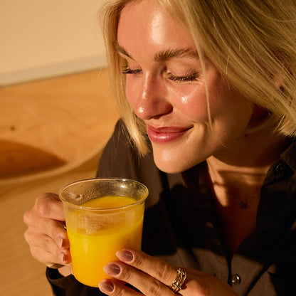 A woman with blonde hair smiles with closed eyes, enjoying a caffeine-free mood boost as she holds a cup of THE FULLEST Saffron Latte in warm, natural light.