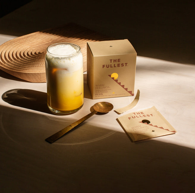 A glass of saffron latte beverage with foam, next to a product box labeled "the fullest" and a spoon, all on a table in sunlight.