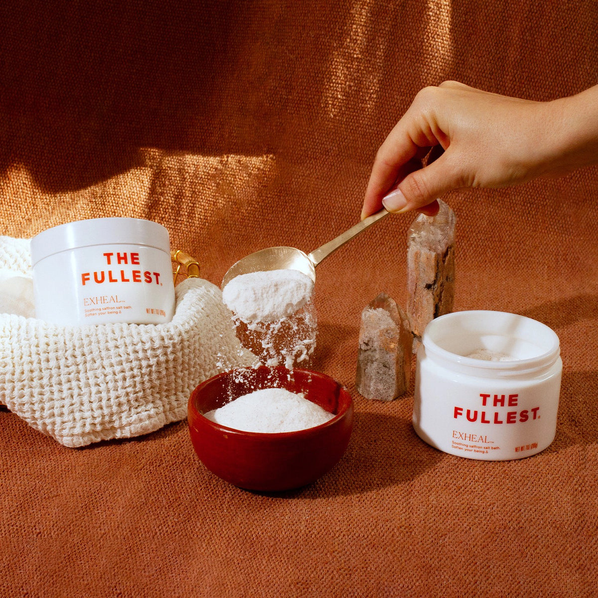 A hand holding a spoon that scoops powder from a white container labeled &quot;Exheal detoxification blend,&quot; with another identical container, a small bowl with powder, and a beige textured background by THE FULLEST.