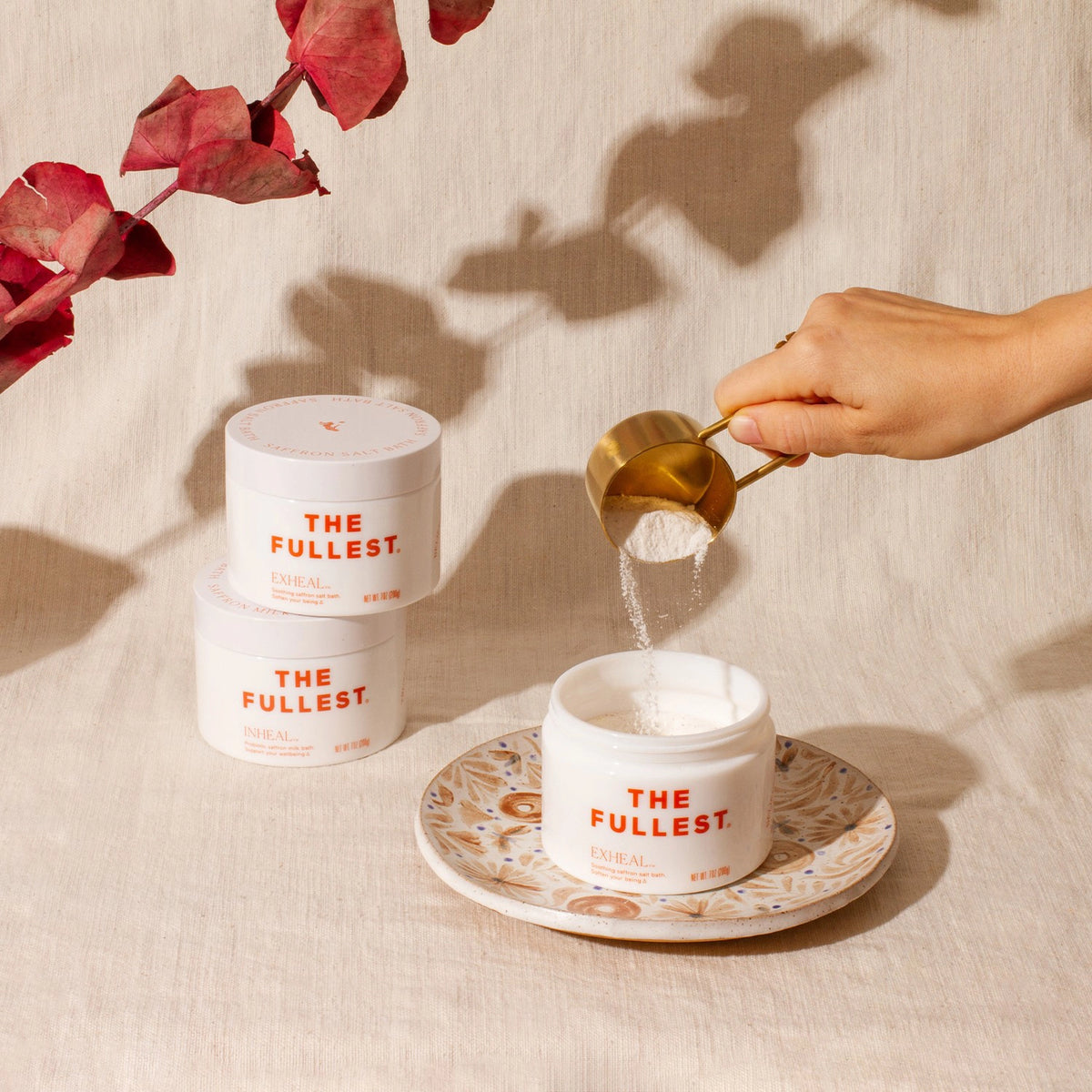 A person&#39;s hand pouring Exheal powder from a brass scoop into a jar labeled &quot;THE FULLEST&quot; on a plate, with two additional jars beside it and shadows of leaves in the background.