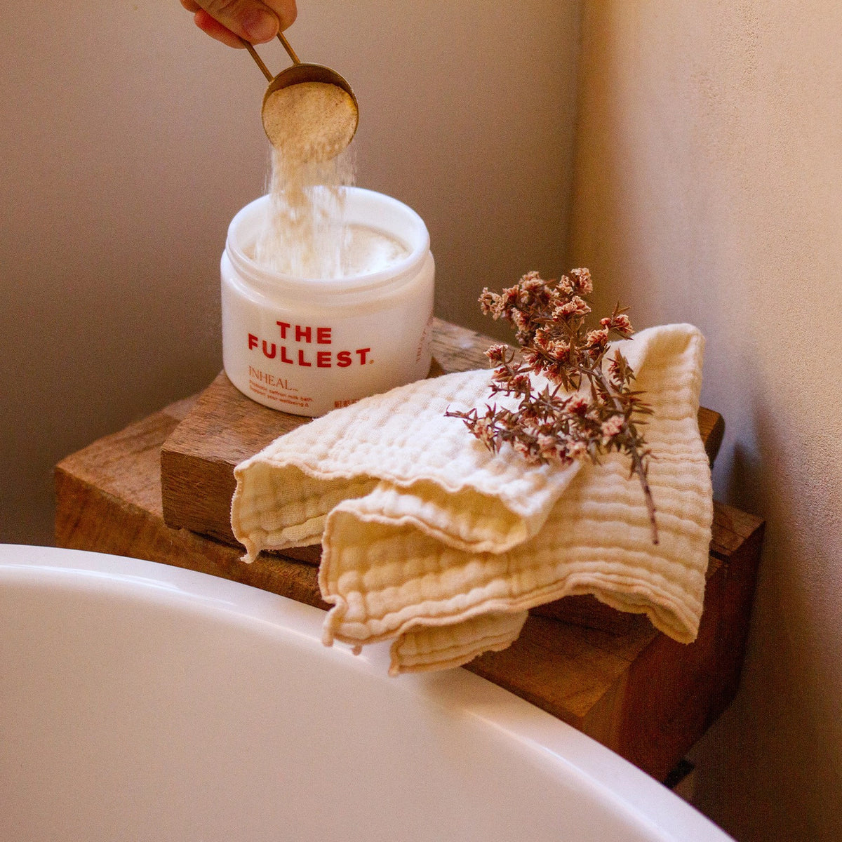 A hand scooping a white powder from a container labeled &quot;Inheal saffron milk bath&quot; next to a cloth and dried flowers on a wooden stool beside a bathtub.