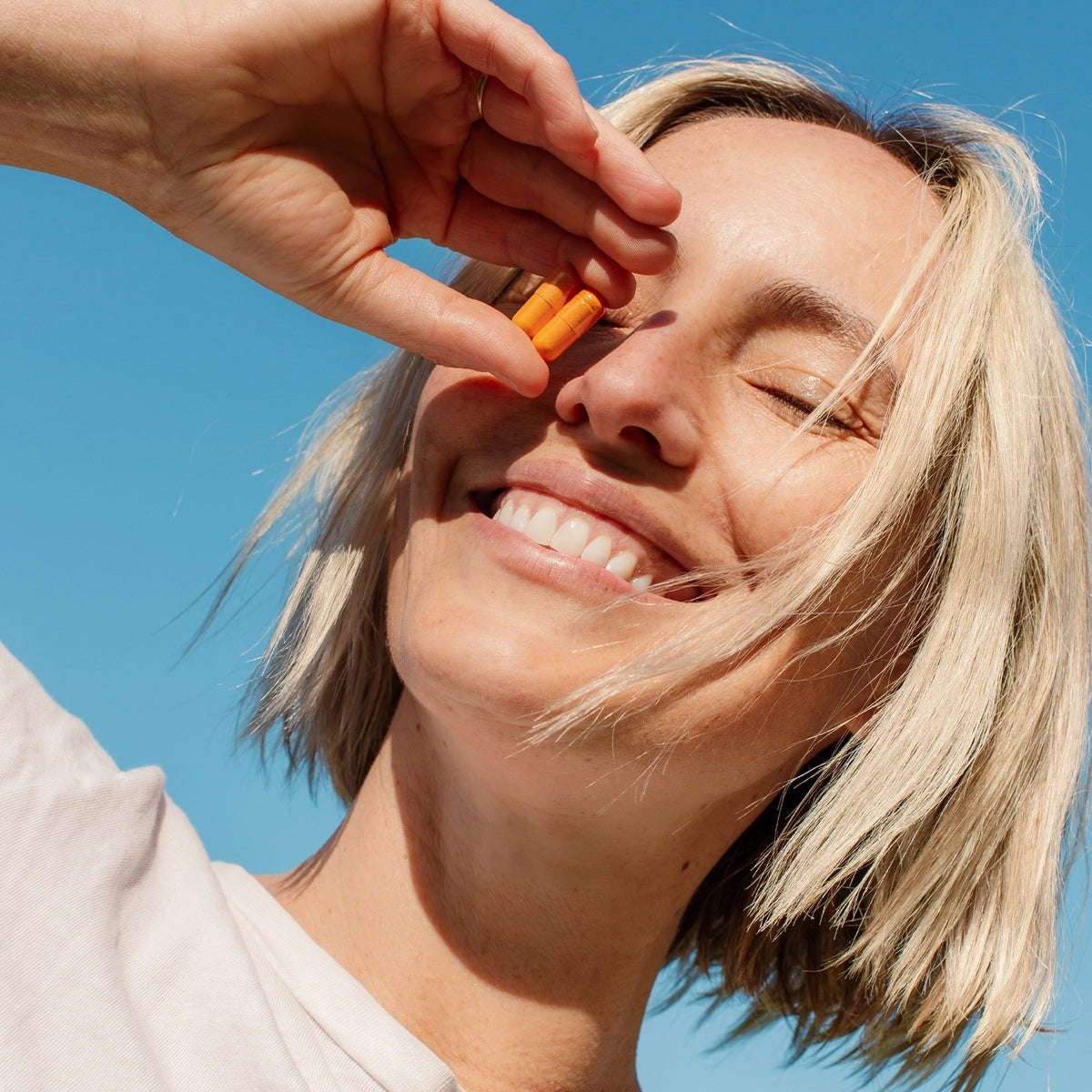 A smiling woman with short blonde hair, holding a Kinder Thoughts supplement capsule between her fingers close to her face, under a clear blue sky.