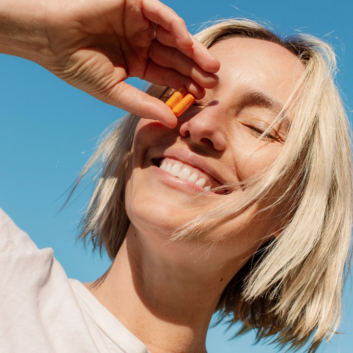 Woman smiling while holding THE FULLEST Kinder Thoughts supplement capsule between her fingers against a clear blue sky.