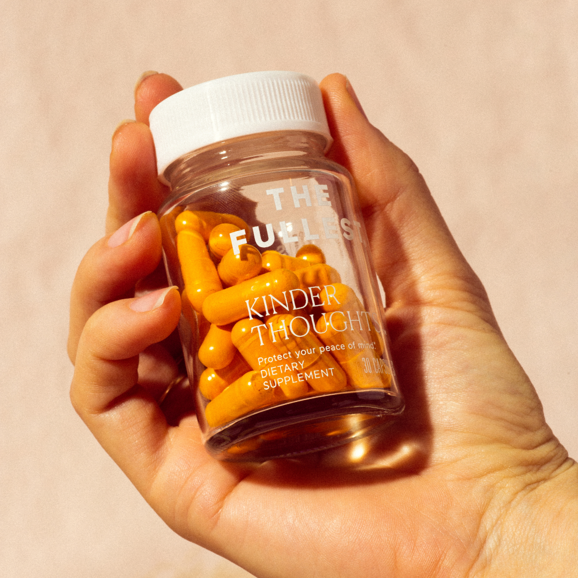 A clear bottle containing orange capsules with a label that reads "Kinder Thoughts," described as a mood-boosting supplement for peace of mind by THE FULLEST.